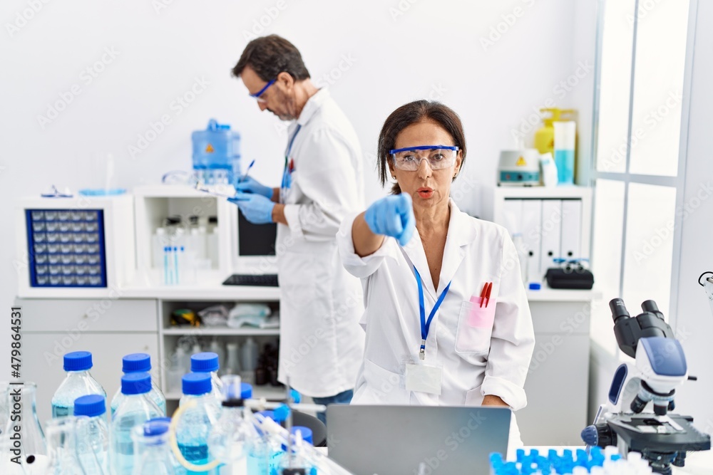 Middle age woman working at scientist laboratory pointing with finger to the camera and to you, confident gesture looking serious