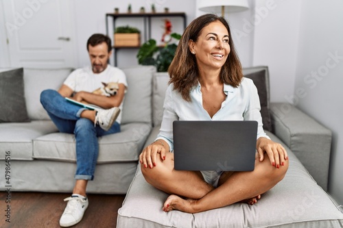 Hispanic middle age couple at home, woman using laptop looking away to side with smile on face, natural expression. laughing confident. © Krakenimages.com