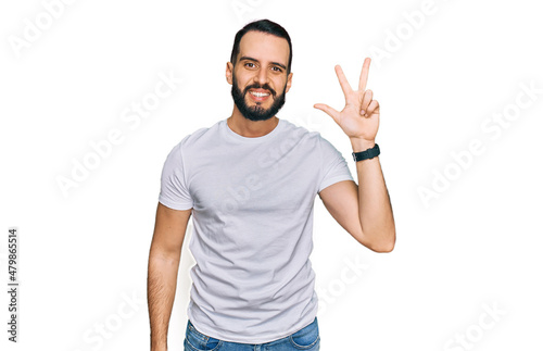 Young man with beard wearing casual white t shirt showing and pointing up with fingers number three while smiling confident and happy.