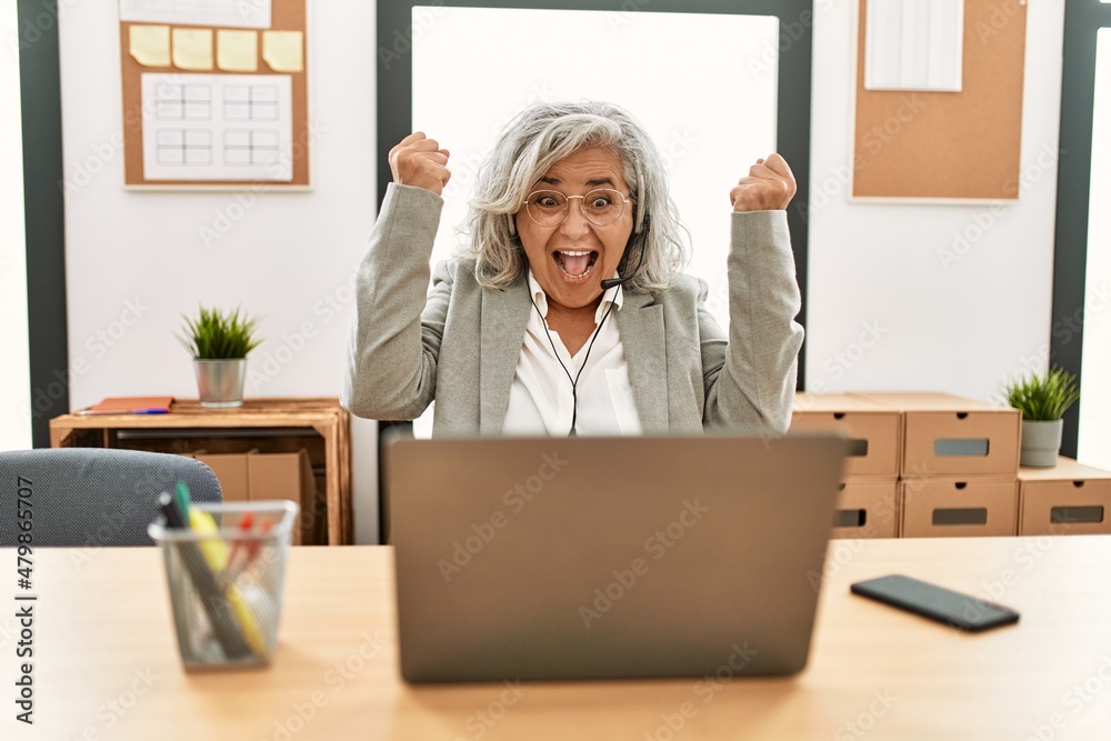 Middle age businesswoman sitting on desk working using laptop at office very happy and excited doing winner gesture with arms raised, smiling and screaming for success. celebration concept. Middle age businesswoman sitting on desk working using laptop at office very happy and excited doing winner gesture with arms raised, smiling and screaming for success. celebration concept.