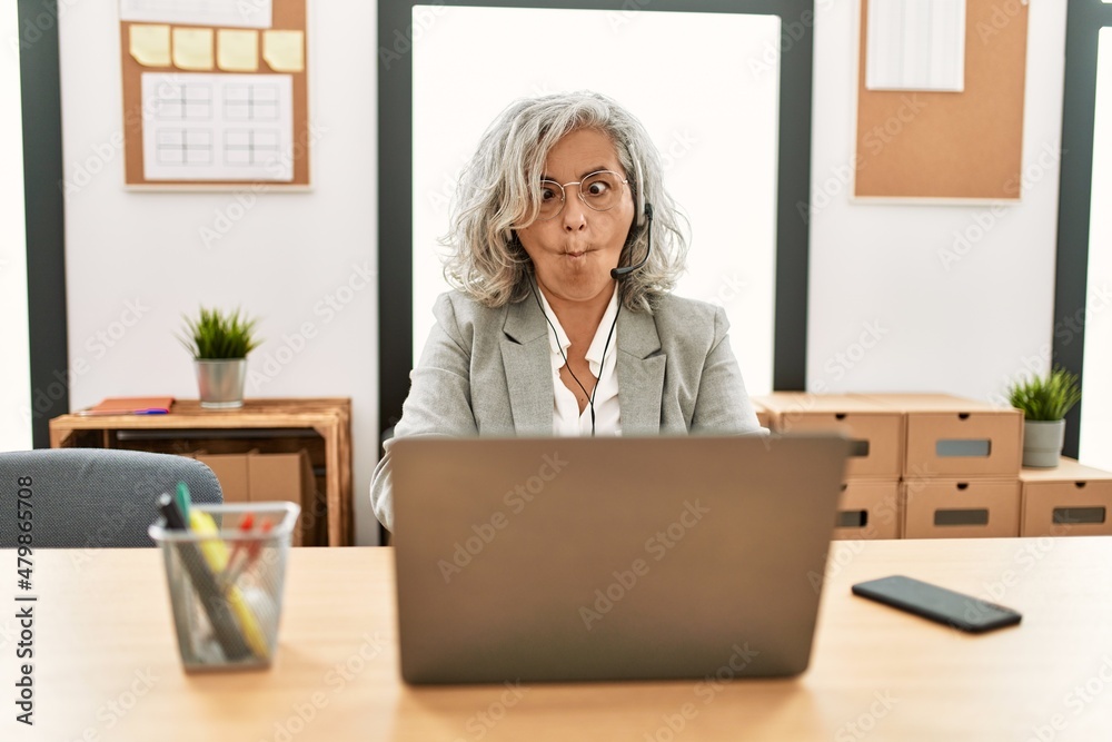 Middle age businesswoman sitting on desk working using laptop at office making fish face with lips, crazy and comical gesture. funny expression.