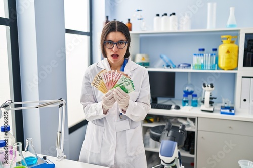 Young hispanic woman working at scientist laboratory holding money banknotes in shock face  looking skeptical and sarcastic  surprised with open mouth