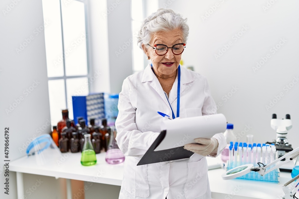 Senior grey-haired woman wearing scientist uniform writing on clipboard at laboratory