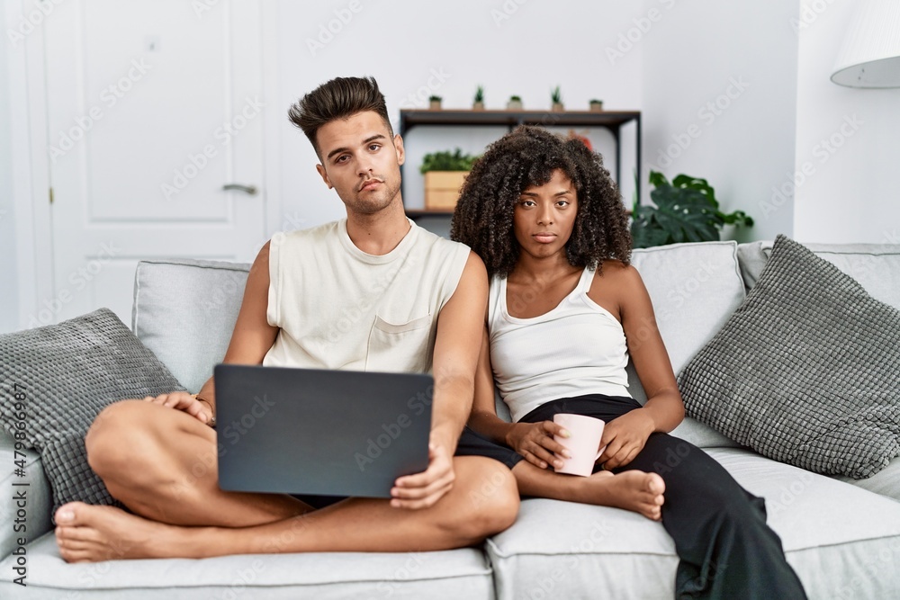 Young interracial couple using laptop at home sitting on the sofa looking sleepy and tired, exhausted for fatigue and hangover, lazy eyes in the morning.