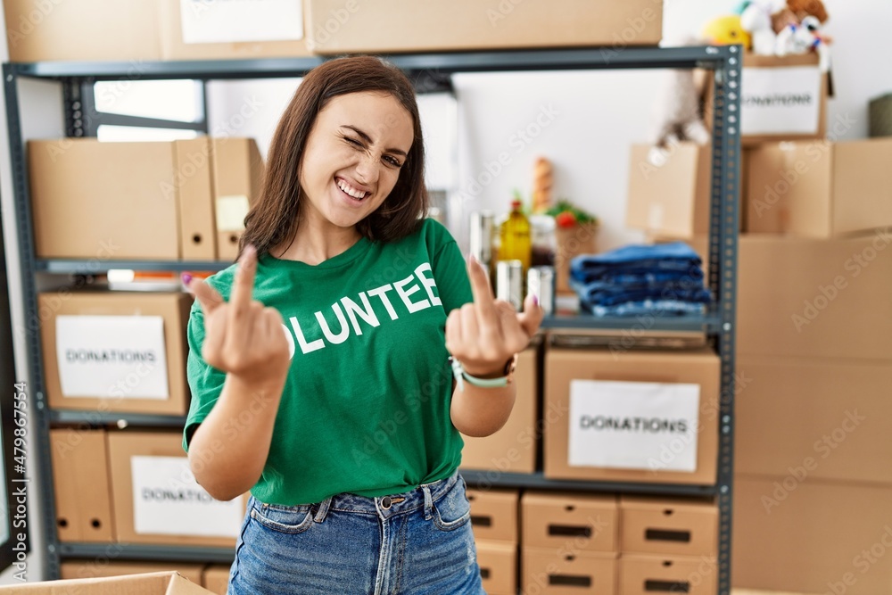 Young brunette woman wearing volunteer t shirt at donations stand showing middle finger doing fuck you bad expression, provocation and rude attitude. screaming excited Young brunette woman wearing volunteer t shirt at donations stand showing middle finger doing fuck you bad expression, provocation and rude attitude. screaming excited