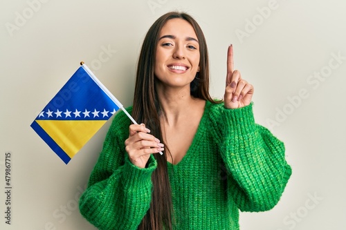 Young hispanic girl holding bosnia herzegovina flag smiling with an idea or question pointing finger with happy face, number one photo