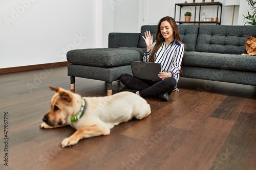 Young hispanic woman having video call sitting on floor with dogs at home