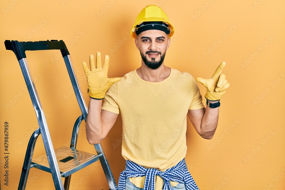 Handsome man with beard by construction stairs wearing hardhat showing and pointing up with fingers number seven while smiling confident and happy. Handsome man with beard by construction stairs wearing hardhat showing and pointing up with fingers number seven while smiling confident and happy.