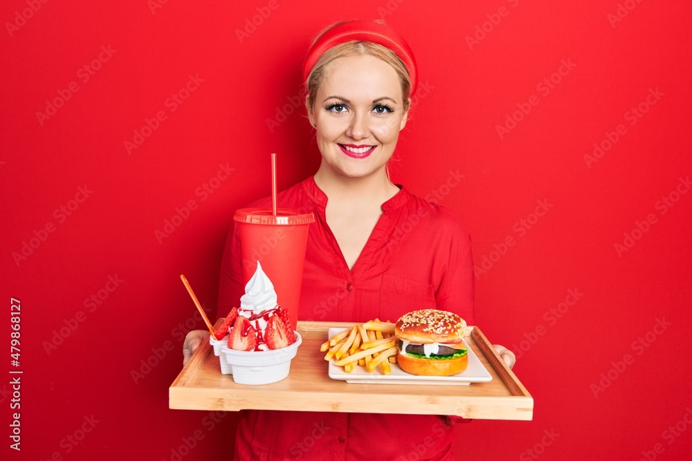 Young blonde woman eating a tasty classic burger with fries and soda smiling with a happy and cool smile on face. showing teeth. Young blonde woman eating a tasty classic burger with fries and soda smiling with a happy and cool smile on face. showing teeth.