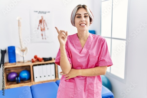 Young nurse woman working at pain recovery clinic smiling happy pointing with hand and finger to the side