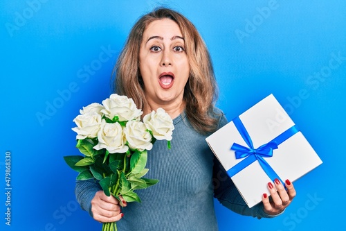 Middle age caucasian woman holding anniversary present and bouquet of flowers afraid and shocked with surprise and amazed expression, fear and excited face.