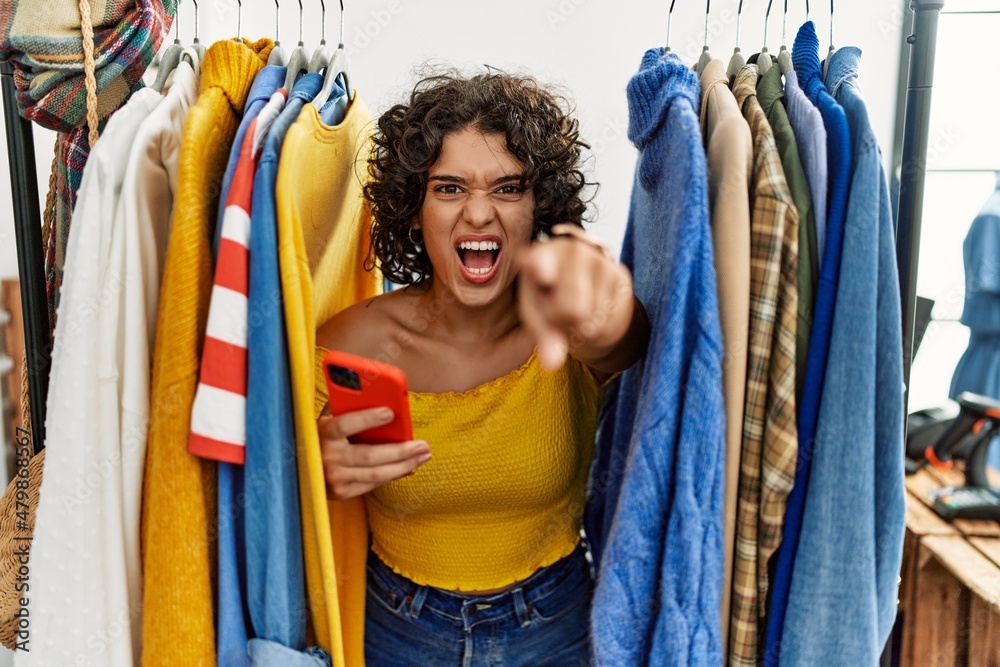 Obraz premium Young hispanic woman searching clothes on clothing rack using smartphone pointing displeased and frustrated to the camera, angry and furious with you