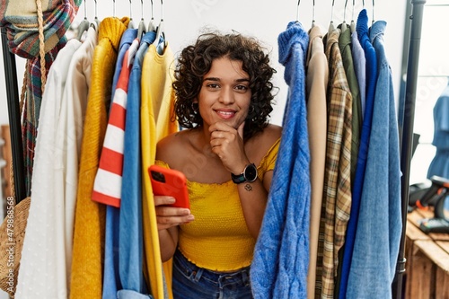 Young hispanic woman searching clothes on clothing rack using smartphone looking confident at the camera smiling with crossed arms and hand raised on chin. thinking positive.
