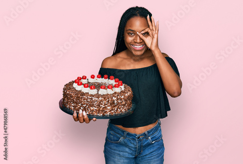Young african american woman celebrating birthday holding big chocolate cake smiling happy doing ok sign with hand on eye looking through fingers