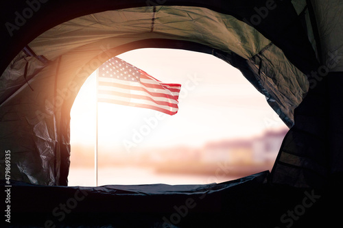 View from a tent of a homeless on a waving flag of USA and down town out of focus in the background at sunrise. Social issue, inadequate affordable housing and income supply concept. Sun flare. photo