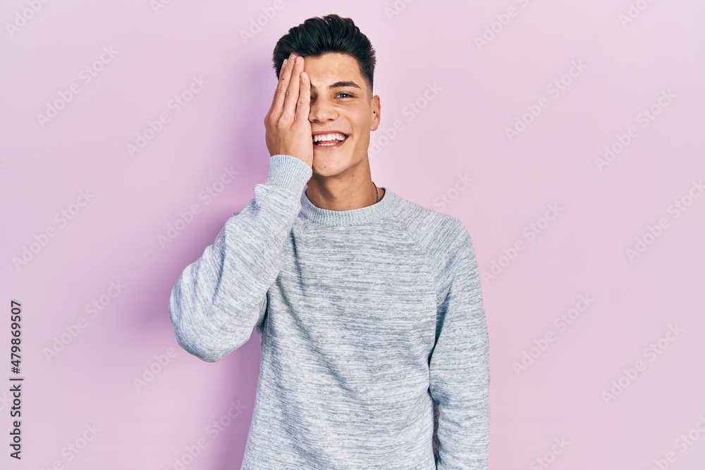Young hispanic man wearing casual clothes covering one eye with hand, confident smile on face and surprise emotion.