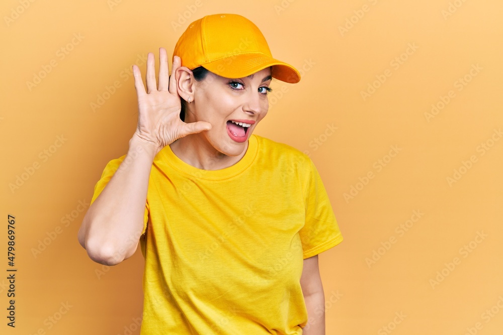 Young hispanic woman wearing delivery uniform and cap smiling with hand over ear listening an hearing to rumor or gossip. deafness concept.