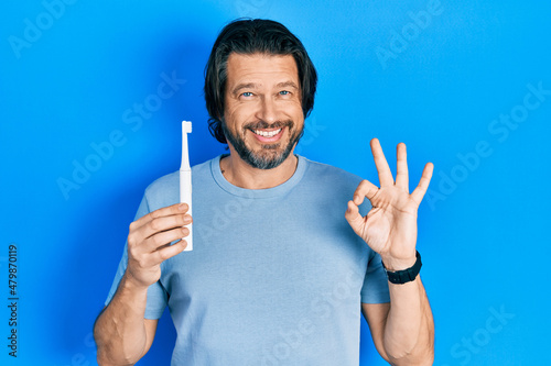 Middle age caucasian man holding electric toothbrush doing ok sign with fingers, smiling friendly gesturing excellent symbol