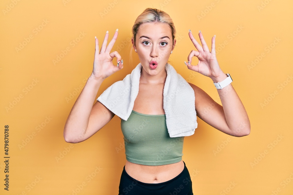 Young blonde girl wearing sportswear and towel looking surprised and shocked doing ok approval symbol with fingers. crazy expression Young blonde girl wearing sportswear and towel looking surprised and shocked doing ok approval symbol with fingers. crazy expression