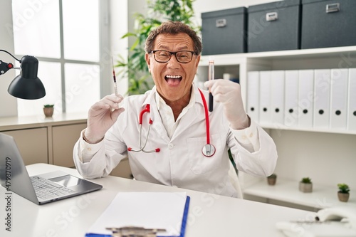 Senior doctor man holding syringe and blood sample smiling and laughing hard out loud because funny crazy joke.