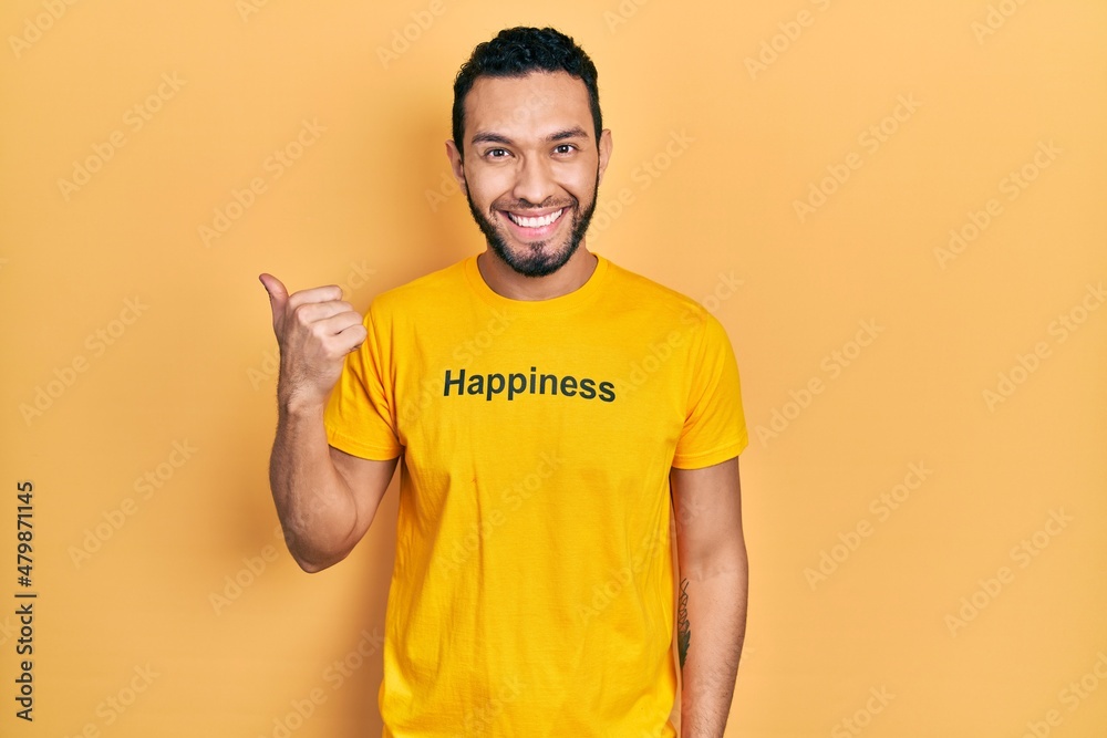 Hispanic man with beard wearing t shirt with happiness word message pointing to the back behind with hand and thumbs up, smiling confident Hispanic man with beard wearing t shirt with happiness word message pointing to the back behind with hand and thumbs up, smiling confident