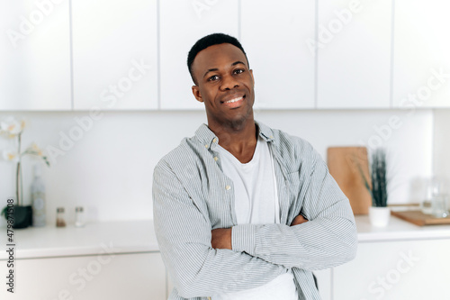 Portrait of African American handsome guy in the apartment. Black attractive man, dressed in stylish casual clothes, stands at home in the kitchen with crossed arms, looks at the camera,smile friendly