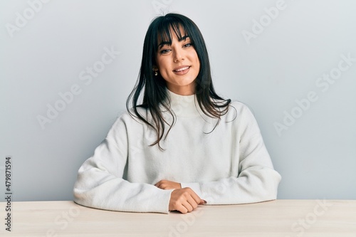 Obraz na plátně Young brunette woman with bangs wearing casual clothes sitting on the table with a happy and cool smile on face