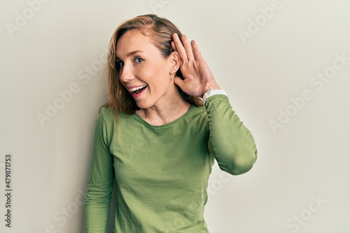 Young blonde woman wearing casual clothes smiling with hand over ear listening and hearing to rumor or gossip. deafness concept.