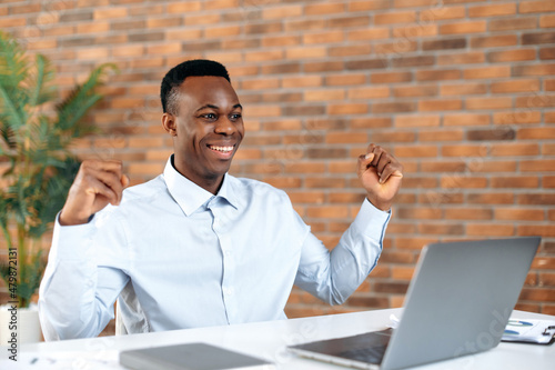 Success, win, luck. African american excited man, broker, agent, manager, working on laptop in modern office, amazed looks at screen, enjoying good deal, big profit, great news, gesturing with fists