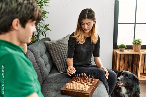 Young caucasian couple smiling happy playing chess game sitting on the sofa with dog at home.