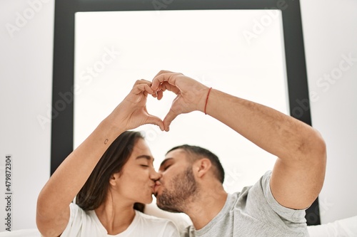 Young hispanic couple doing heart symbol with hands sitting on the bed at home.
