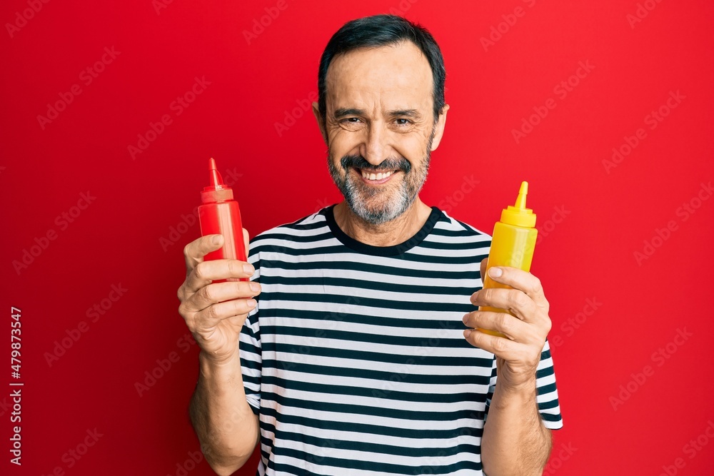 Middle age hispanic man holding ketchup and mustard bottle smiling with a happy and cool smile on face. showing teeth. Middle age hispanic man holding ketchup and mustard bottle smiling with a happy and cool smile on face. showing teeth.