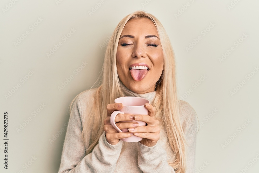 Young blonde woman drinking a cup of coffee sticking tongue out happy with funny expression. Young blonde woman drinking a cup of coffee sticking tongue out happy with funny expression.