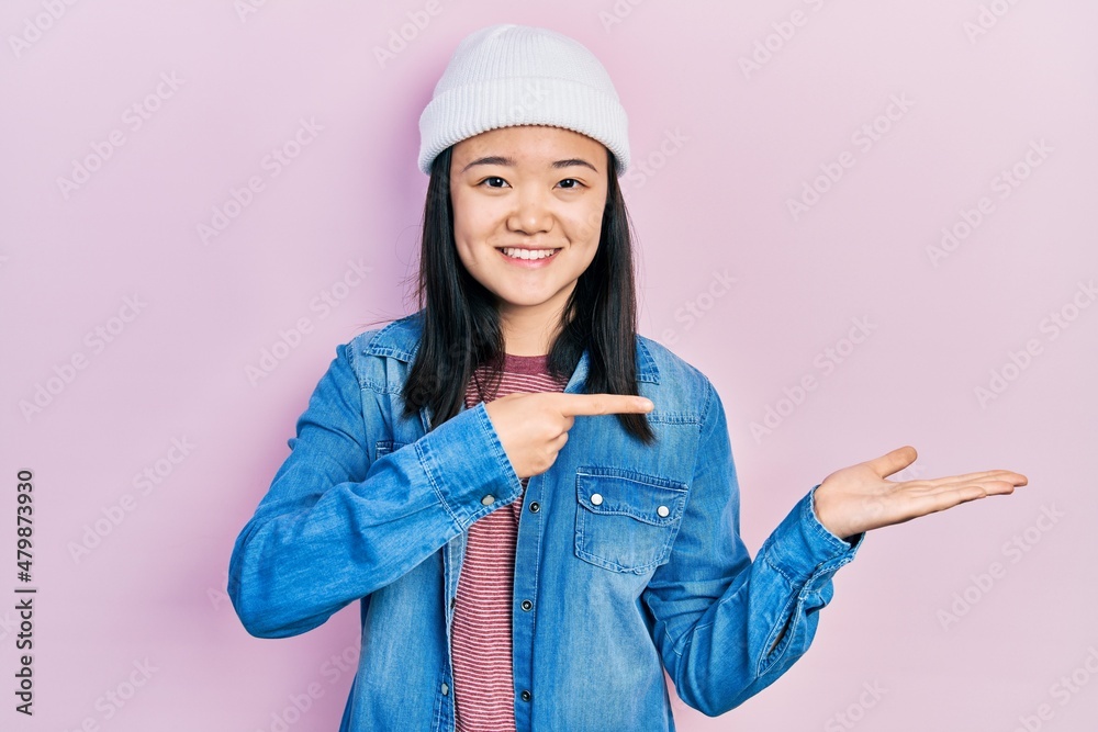 Young chinese girl wearing cute wool cap amazed and smiling to the camera while presenting with hand and pointing with finger. Young chinese girl wearing cute wool cap amazed and smiling to the camera while presenting with hand and pointing with finger.