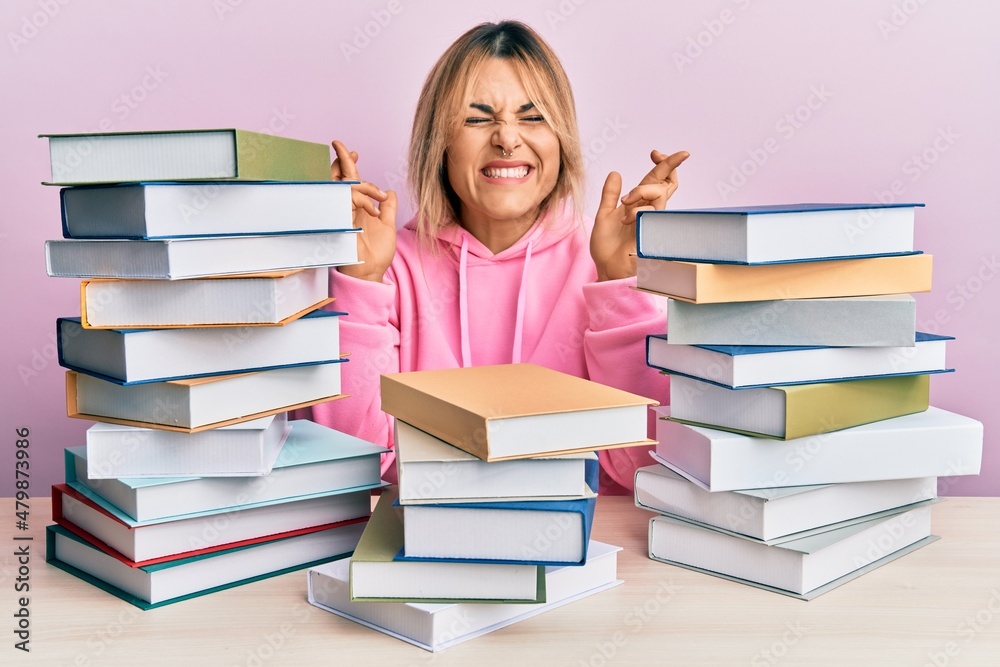 Young caucasian woman sitting on the table with books gesturing finger crossed smiling with hope and eyes closed. luck and superstitious concept. Young caucasian woman sitting on the table with books gesturing finger crossed smiling with hope and eyes closed. luck and superstitious concept.