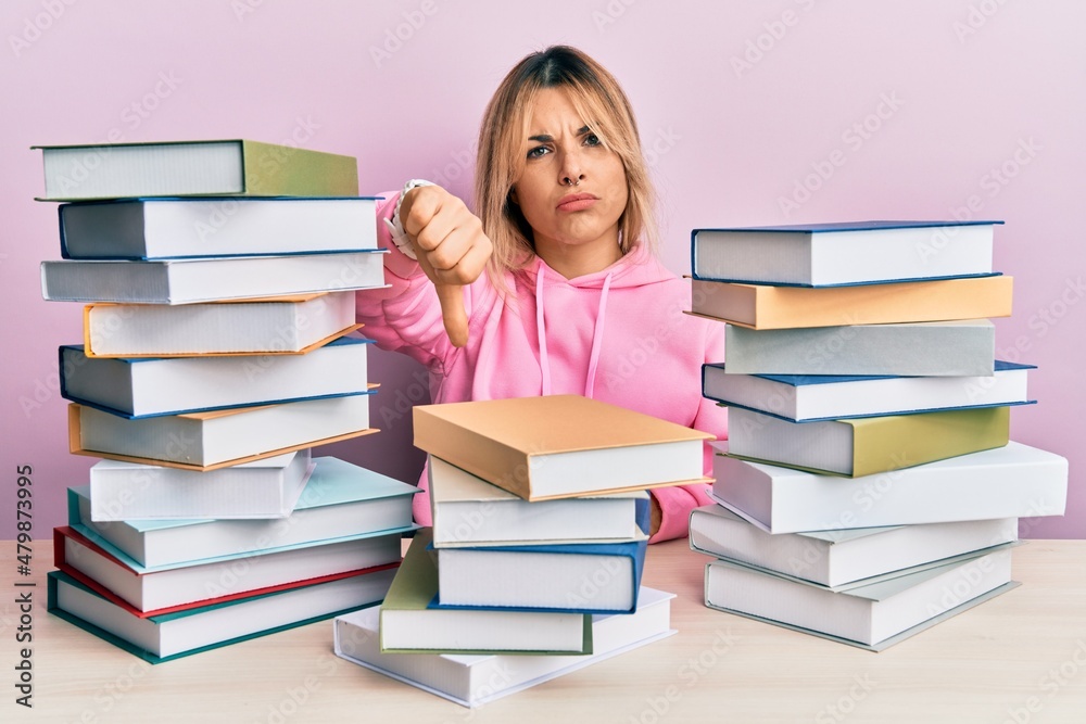Young caucasian woman sitting on the table with books looking unhappy and angry showing rejection and negative with thumbs down gesture. bad expression. Young caucasian woman sitting on the table with books looking unhappy and angry showing rejection and negative with thumbs down gesture. bad expression.