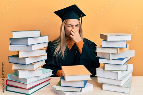 Young caucasian woman wearing graduation ceremony robe sitting on the table smelling something stinky and disgusting, intolerable smell, holding breath with fingers on nose. bad smell
