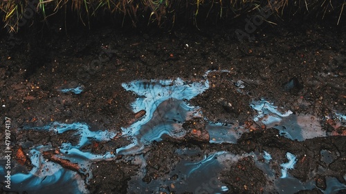 Smudges of Toxic Industrial Liquid Chemical Pollutant Visible on Surface of Rain Water Puddles on Muddy Ground Road	