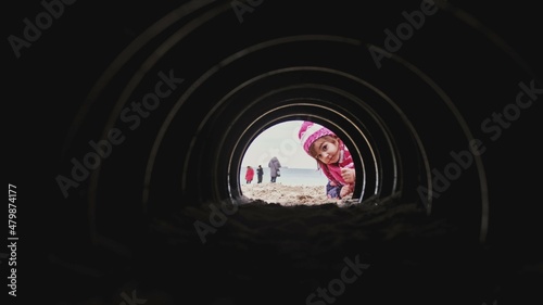 Fototapeta Naklejka Na Ścianę i Meble -  Curious Caucasian Girl in Warm Clothes Looking and Crawling into Dark Narrow Tunnel on Beach	