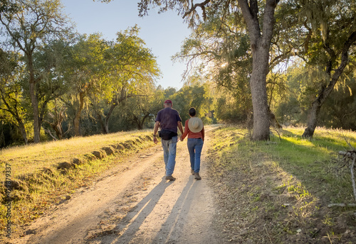 Male and female couple walking together holding hands on romantic stroll on dirt path into the sunset through the oak trees, casting long shadows  © Dennis M. Swanson