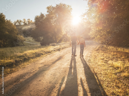 Romantic male and female couple walking hand-in-hand on dirt road with sunlight flooding through trees. Couple in romantic stroll into sunset.
