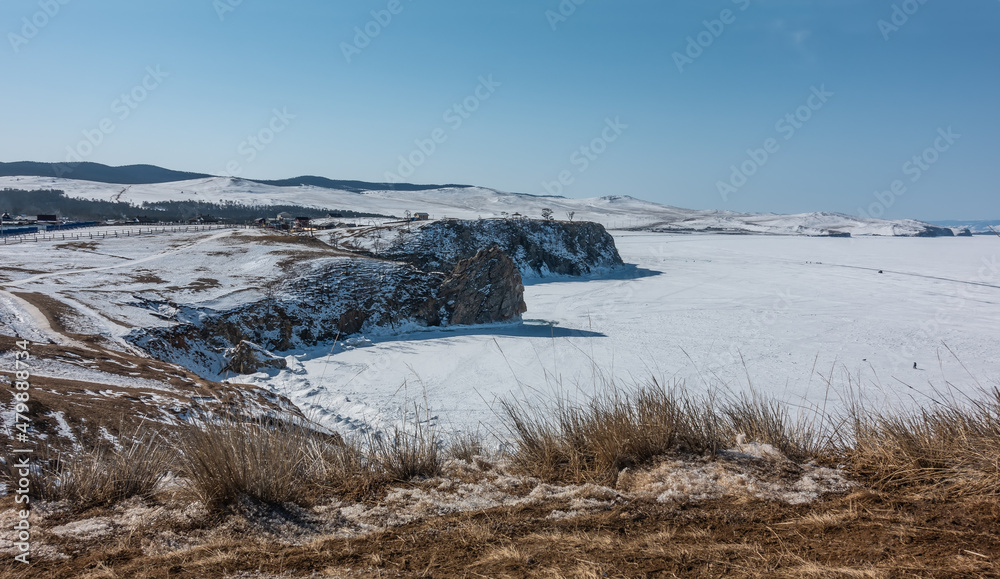 The steep shores of the frozen lake are covered with snow. Village houses are visible in the distance. Clear blue sky. There is dry grass in the foreground. Baikal The steep shores of the frozen lake are covered with snow. Village houses are visible in the distance. Clear blue sky. There is dry grass in the foreground. Baikal