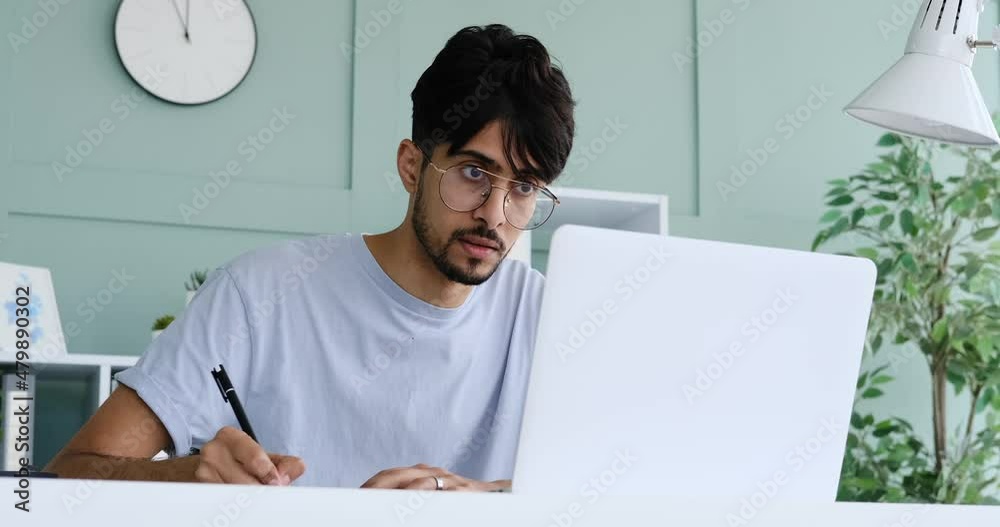 Businessman writing notes while discussing on video call using laptop at office