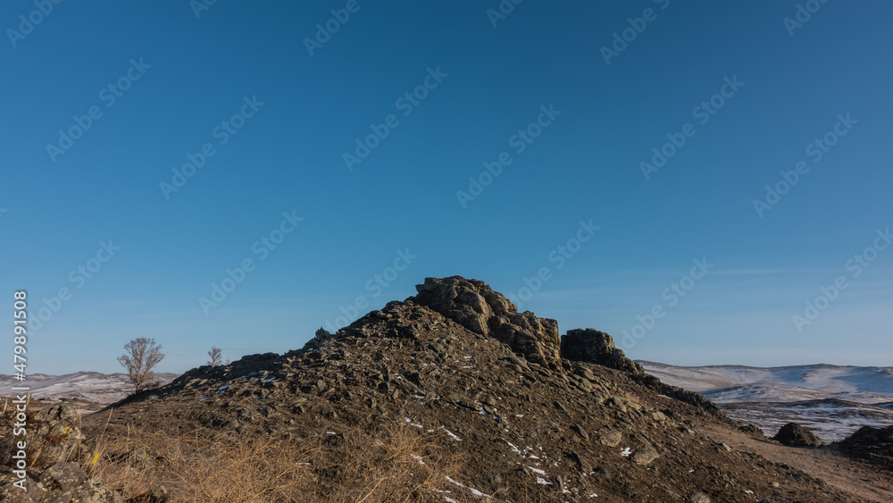 A rocky hill against a clear blue sky. A snow-covered mountain range in the distance. Dry grass and bare trees in the foreground. Siberia. Copy Space