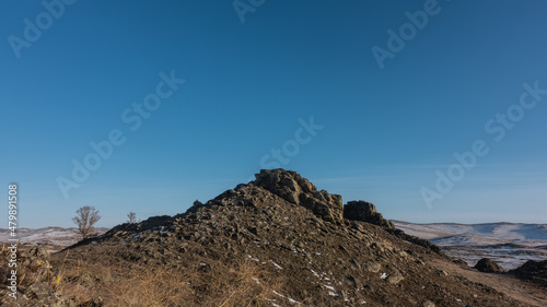 A rocky hill against a clear blue sky. A snow-covered mountain range in the distance. Dry grass and bare trees in the foreground. Siberia. Copy Space