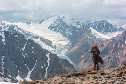 Hiker at very high altitude with view to long glacier tongue and high snow mountains under cloudy sky. Man on windy stone hill against huge mountains with low clouds. Man and majestic mountains nature photo