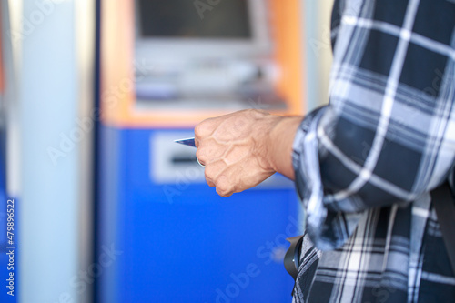 A male tourist in a long-sleeved striped shirt with a camera stands in front of an ATM, happily waiting to withdraw cash for a long weekend. © Tanawit