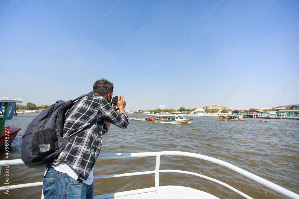 A South Asian tourist, wearing a striped shirt, picked up a camera to photograph Wat Arun on the ferry. and a backpack to walk happily during the long holiday happily in Bangkok, Thailand