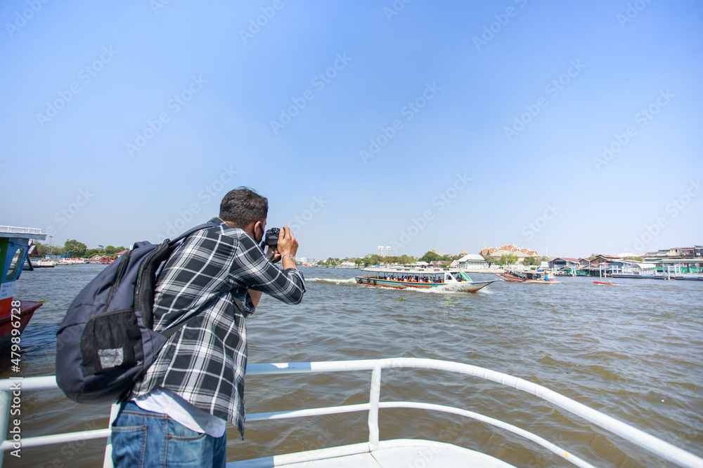 Obraz premium A South Asian tourist, wearing a striped shirt, picked up a camera to photograph Wat Arun on the ferry. and a backpack to walk happily during the long holiday happily in Bangkok, Thailand