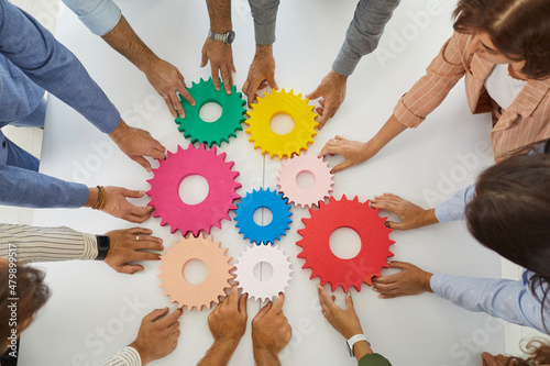 Team of young and senior business people holding and joining colorful gear wheels on office desk as symbol or metaphor for teamwork efficiency and effective collaboration. Top overhead view from above